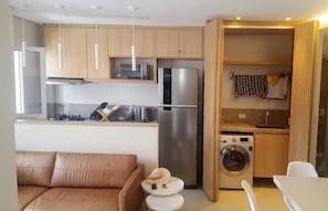 A compact kitchen with a stainless steel refrigerator, a microwave, and a washing machine. There is a wooden cabinet with a microwave above it, and a small table with a chair in the foreground.