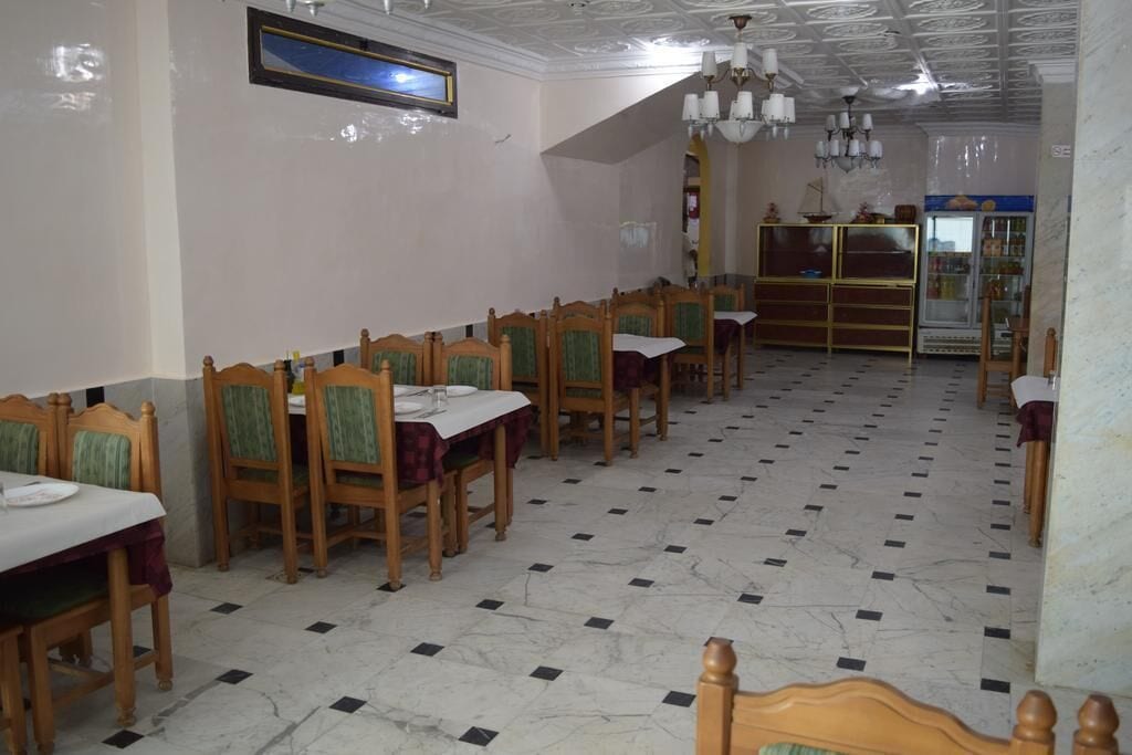 Zona pranzo con sedie e tavoli di legno, un lampadario e un frigorifero sullo sfondo.