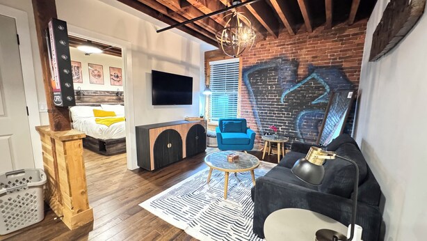 A modern living room with a brick wall, wooden ceiling beams, a large TV, a blue sofa, a round coffee table, and a patterned rug.