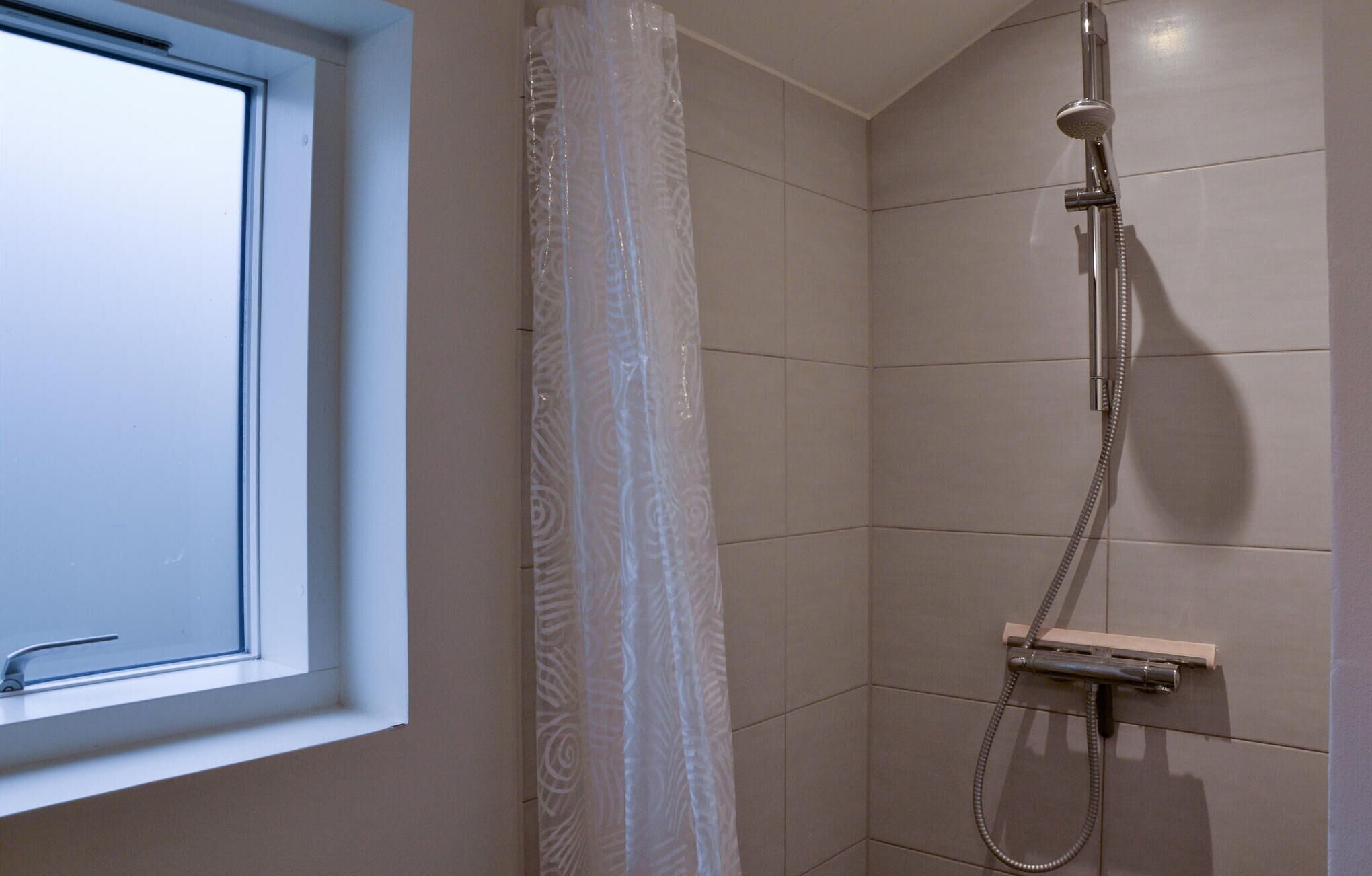A shower with a handheld showerhead and a wall-mounted control panel, a window with a white frame, and a patterned shower curtain.