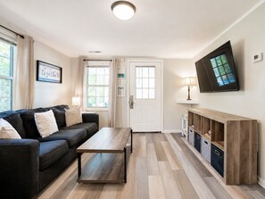 A living room with a sofa, a coffee table, a TV mounted on the wall, and a door with a window. - Cozy Cottage-Screened Porch-Central Location -Beach Access Across- Pet Considered (Folly Beach)