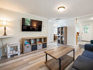 A living room with a flat-screen TV, wooden shelving, a coffee table, and a sofa. - Cozy Cottage-Screened Porch-Central Location -Beach Access Across- Pet Considered (Folly Beach)