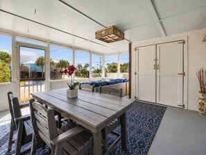 A room with a wooden table, chairs, a blue patterned rug, and a view of the outdoors through large windows. - Cozy Cottage-Screened Porch-Central Location -Beach Access Across- Pet Considered (Folly Beach)