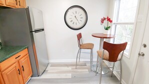Cocina compacta con refrigerador, armarios de madera, una mesa de comedor con dos sillas y un reloj de pared.
