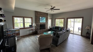 A spacious living room with a fireplace, a ceiling fan, and large windows.