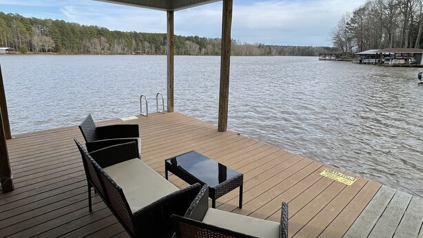 A covered deck with outdoor furniture, overlooking a lake with boats and a dock in the background.