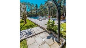 Un espace aménagé au bord de la piscine, avec des chaises longues et une table en bois, entouré d’arbres et surplombé d’un ciel bleu clair.