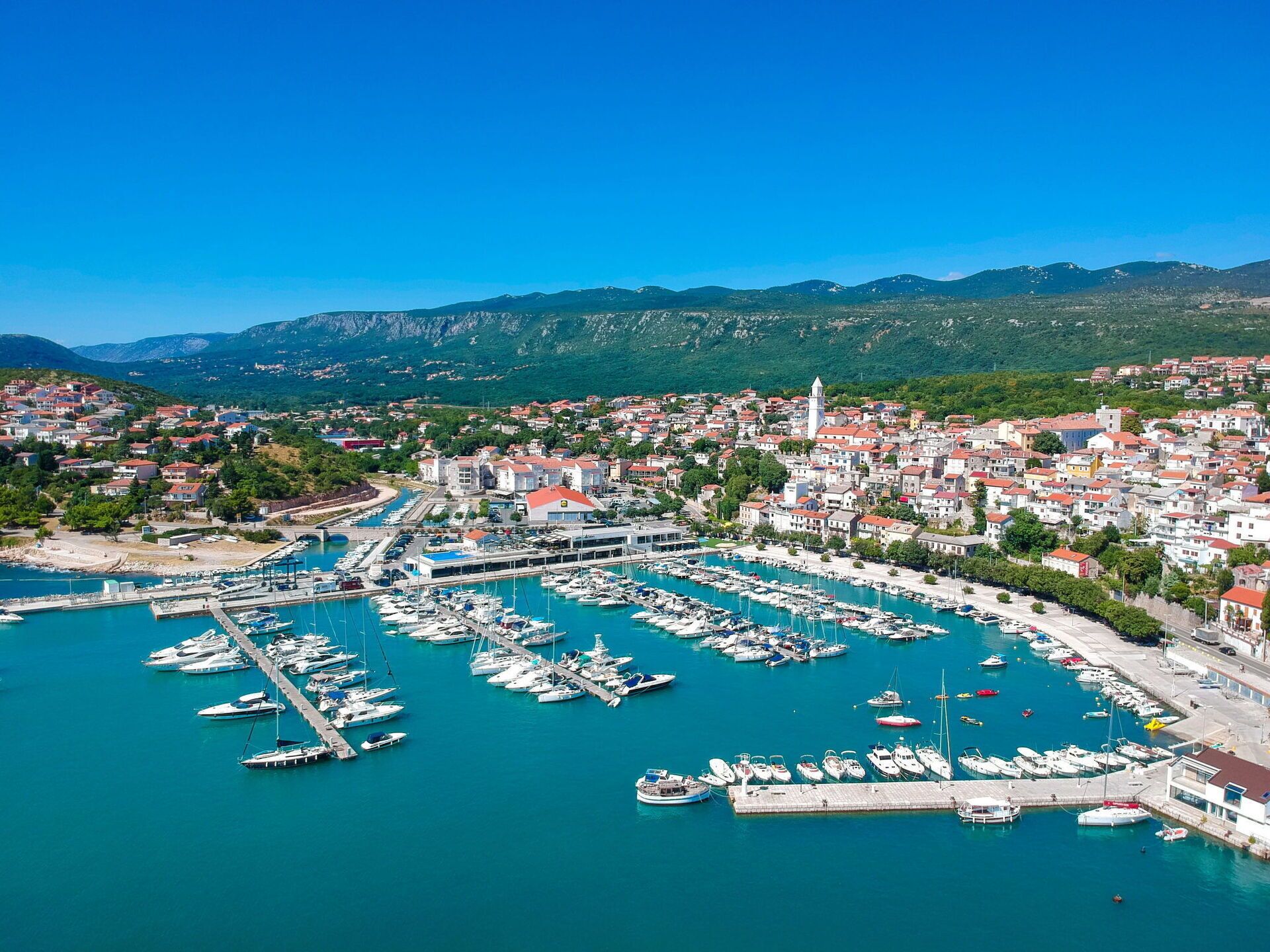 A coastal town with a marina filled with boats, surrounded by residential buildings and green hills in the background.