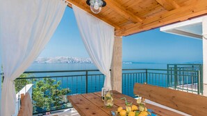 Une terrasse en bois avec une table dressée pour un repas, offrant une vue sur une étendue d’eau et une côte lointaine.