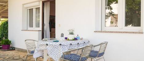 Un patio al aire libre con una mesa blanca cubierta con un mantel de lunares, cuatro sillas de metal con cojines azules y una planta en maceta.