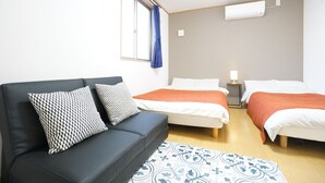 A room with a black sofa, two beds with white and orange bedding, a patterned rug, and a window with blue curtains. - Terry's Apartment Namba South V C01A (Osaka)