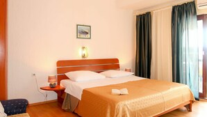 Une chambre d’hôtel avec un lit en bois, une table de chevet avec une lampe, un luminaire fixé au mur, une fenêtre avec des rideaux et un petit tableau accroché au mur.