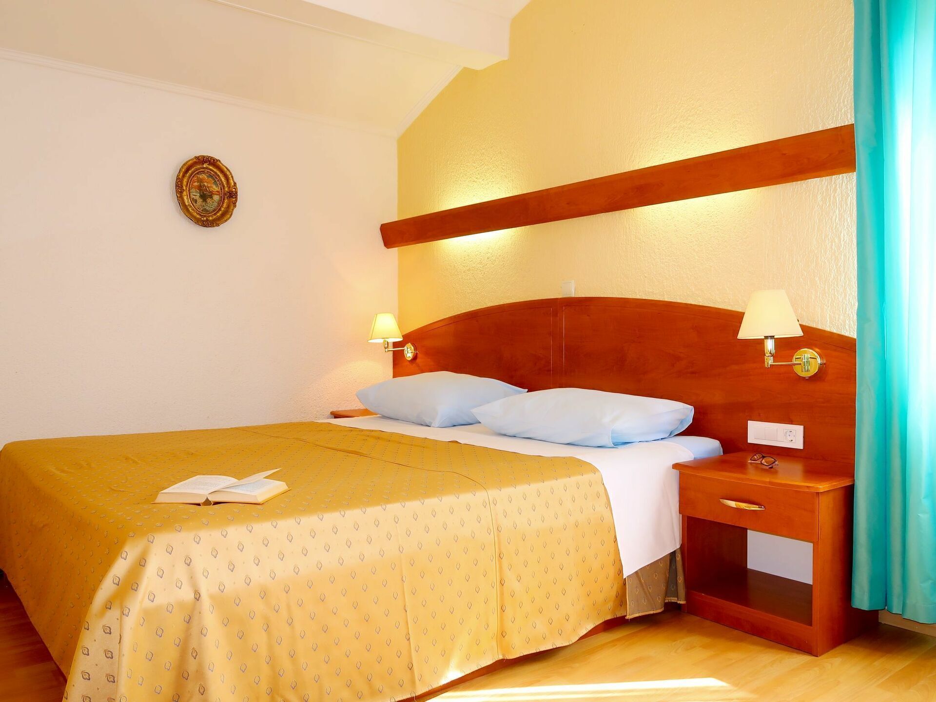 A hotel room with a wooden headboard, two bedside lamps, a bed with a yellow bedspread, and a small wooden nightstand with a drawer.