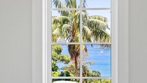 Habitación con una ventana grande que da a la costa, con palmeras y un barco.