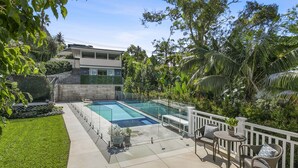 Un área de piscina exterior moderna con barandilla de vidrio, rodeada de exuberante vegetación y una cerca blanca.