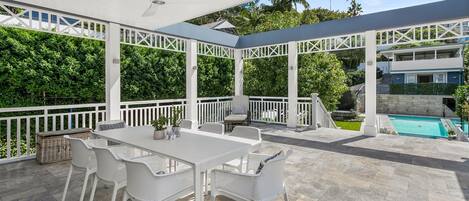 An outdoor patio with a white table and chairs, a ceiling fan, and a view of a swimming pool and greenery.
