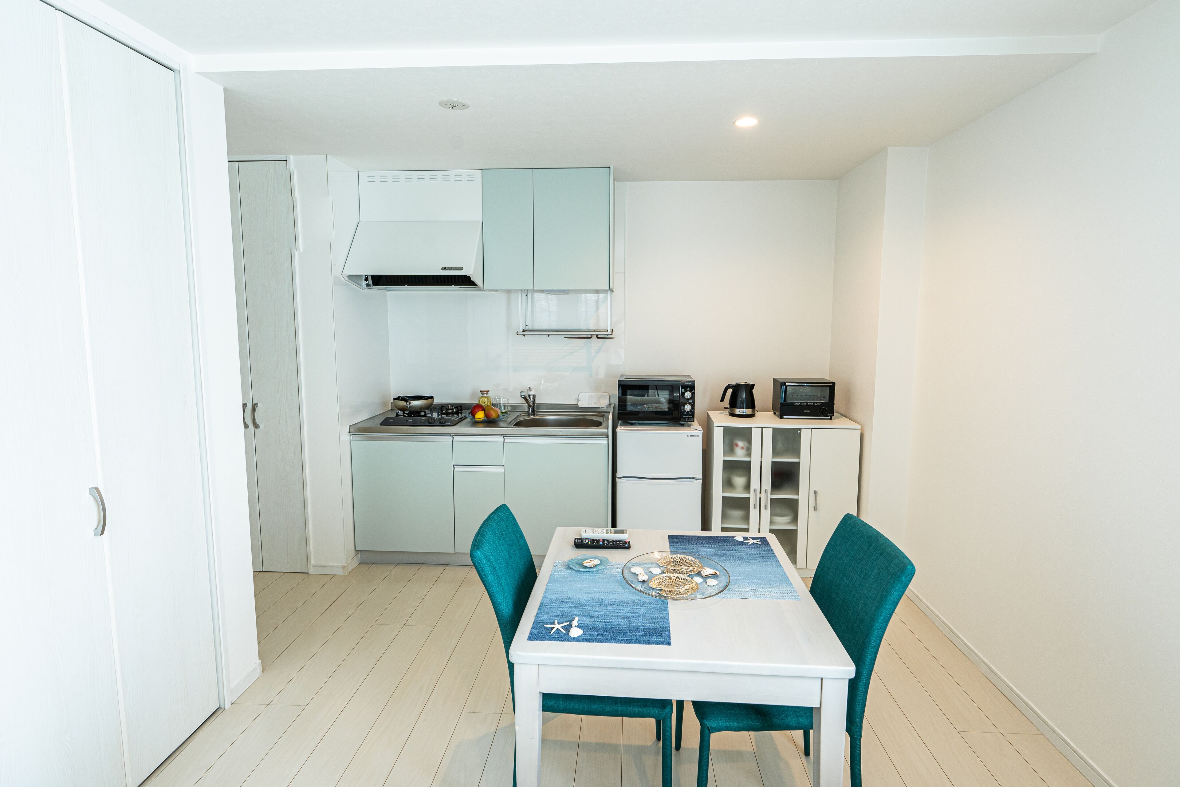 Een compacte keuken met witte kastjes, een magnetron en koffiezetapparaat. Er staat een eettafel met stoelen en een blauwe placemat.