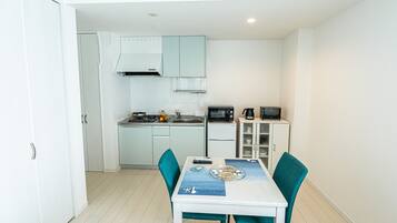 Een compacte keuken met witte kastjes, een magnetron en koffiezetapparaat. Er staat een eettafel met stoelen en een blauwe placemat.