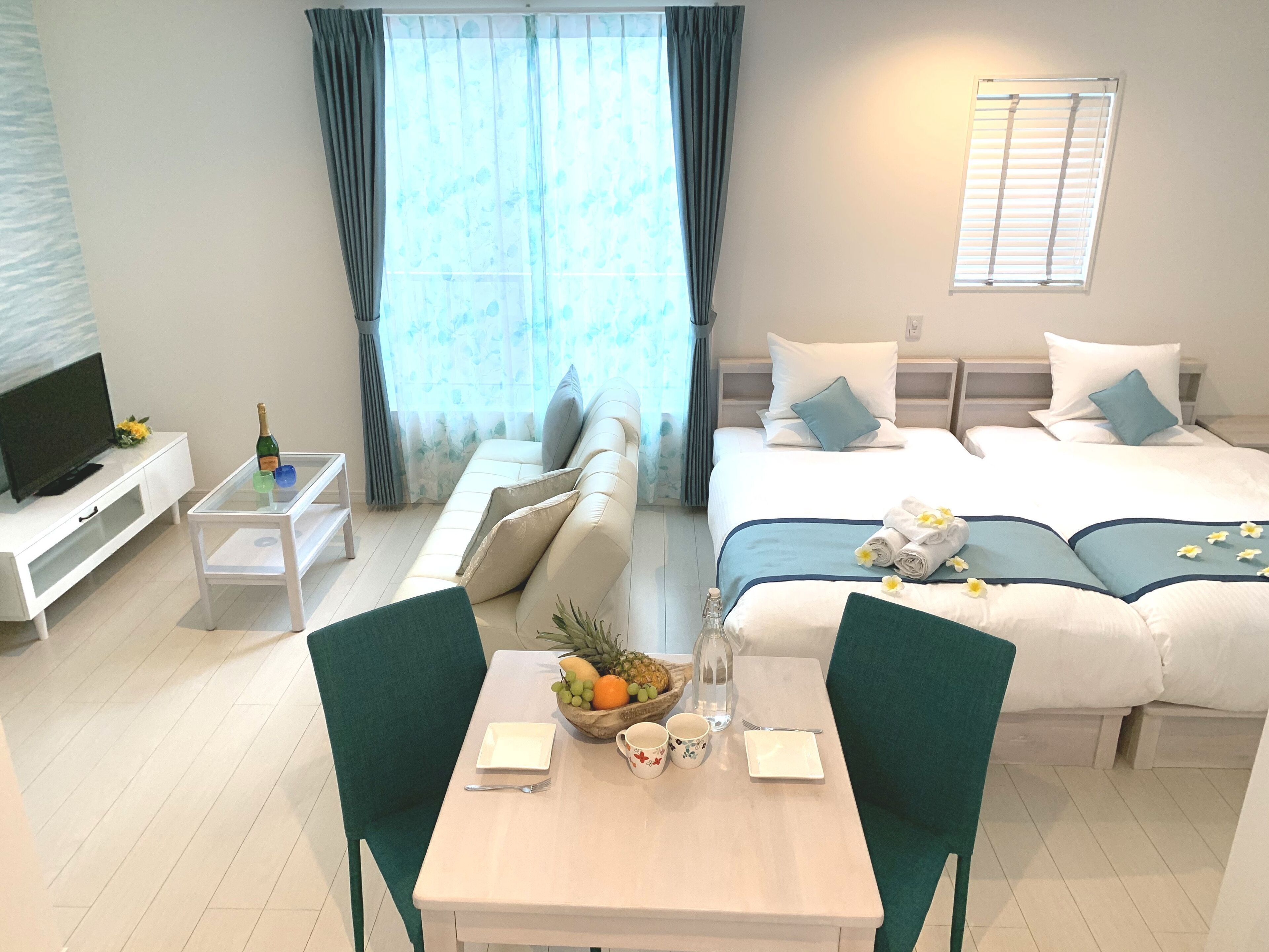 A hotel room with a sofa, a bed with blue bedding, a dining table with chairs, a TV, and a window with curtains.