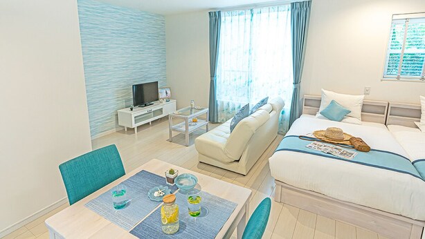 A modern hotel room with a bed, a sofa, a dining table with chairs, a TV, and a window with curtains. - Ecot Kugai 1 (Miyakojima)