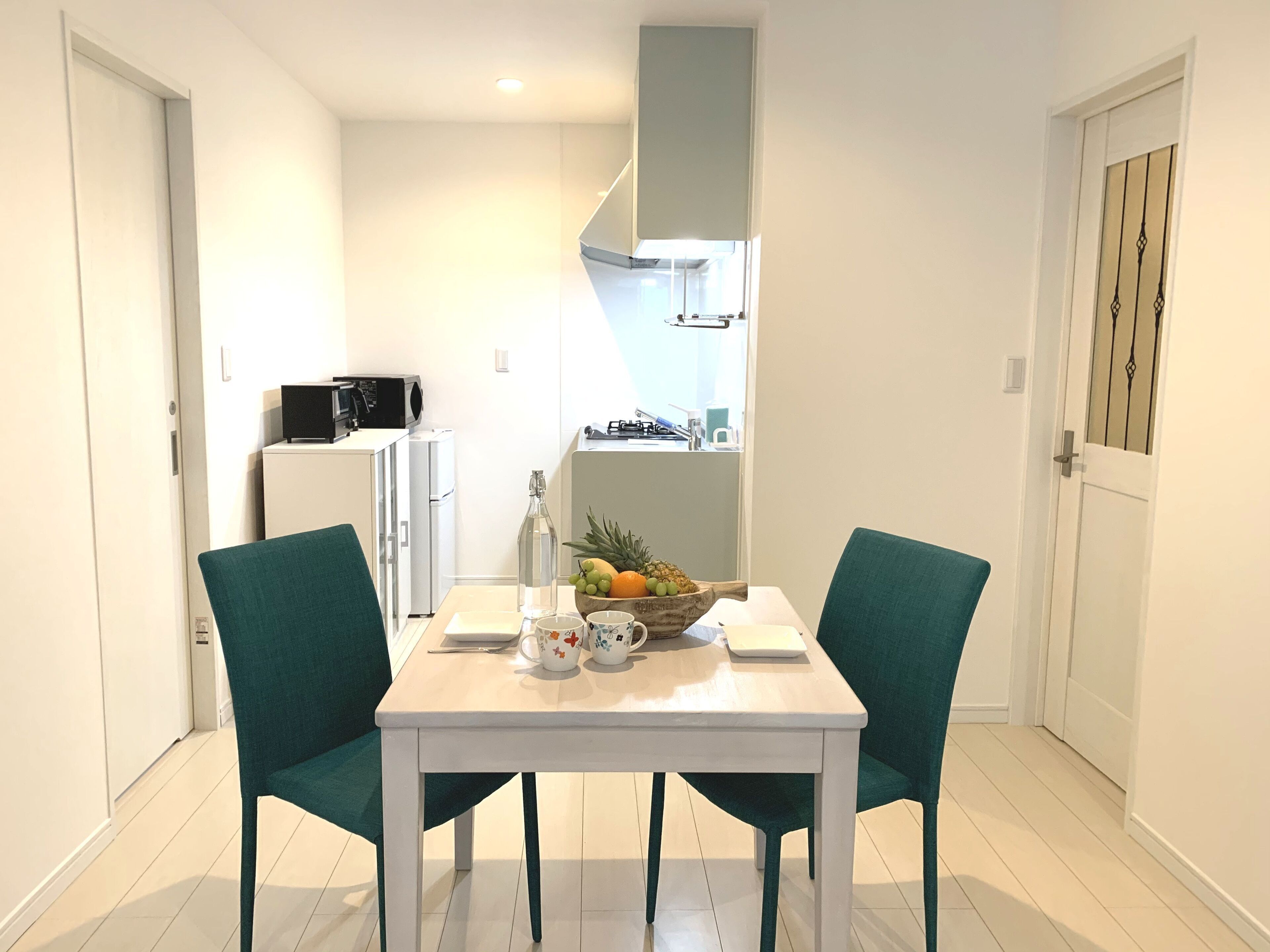 Una habitación pequeña y luminosa con una mesa blanca, dos sillas color turquesa, un refrigerador blanco, un microondas, un lavamanos y una ventana con vistas al cielo.
