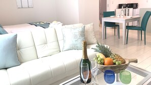 A modern living room with a white sofa, a dining area with a table and chairs, and a kitchen in the background. - Ecot Kugai 1 (Miyakojima)