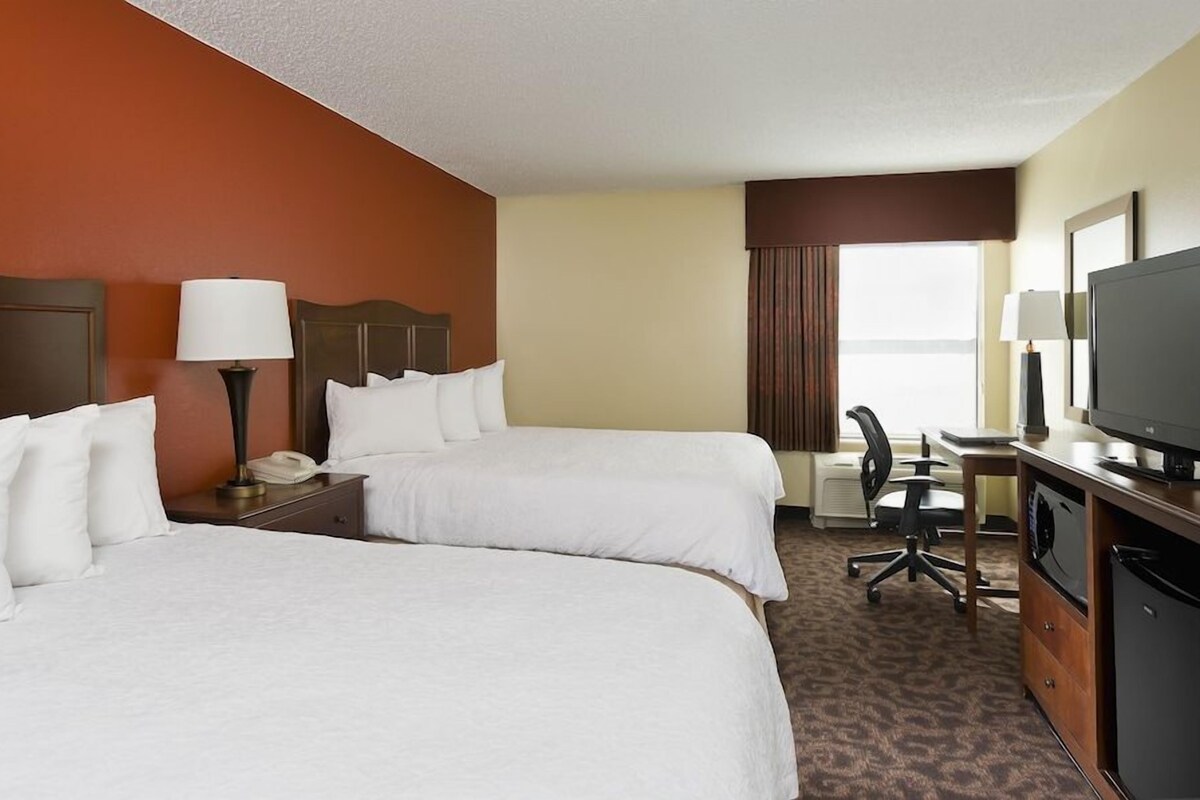 A hotel room with two beds, a desk, a television, and a window with curtains.