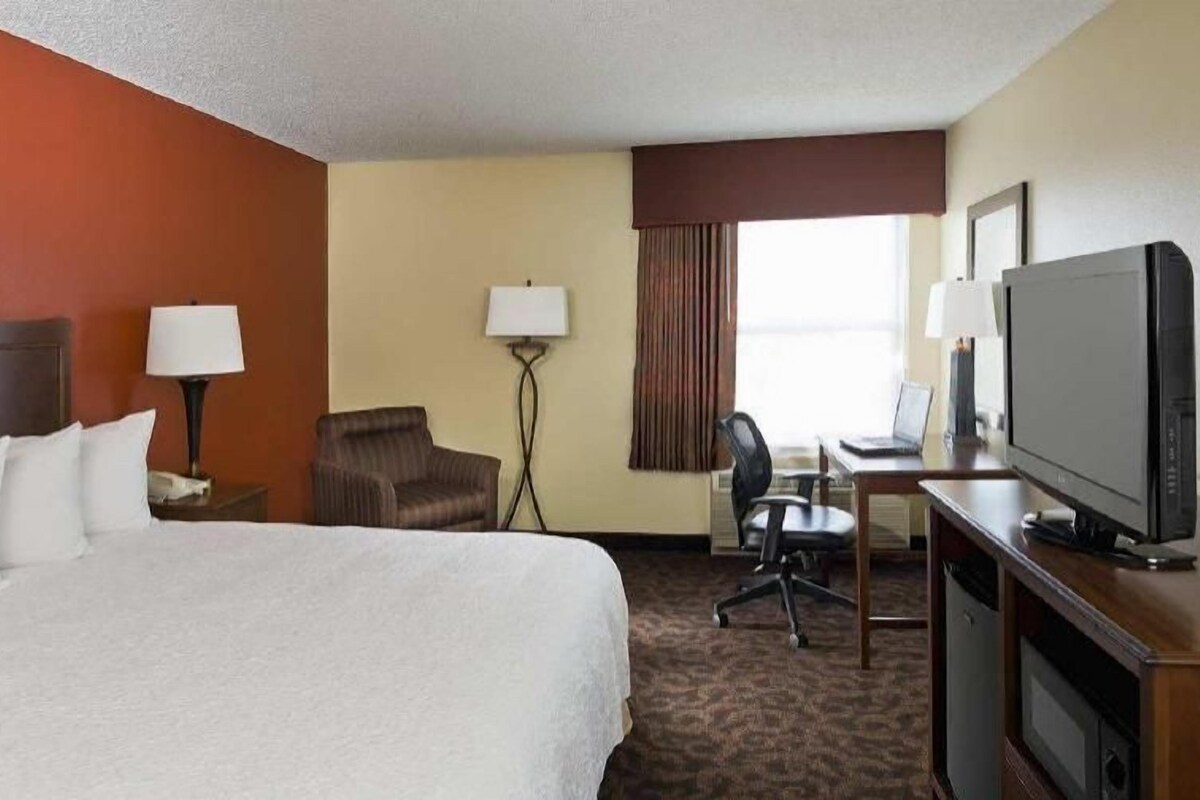 A hotel room with a bed, a desk with a computer, a TV, a chair, and a lamp.