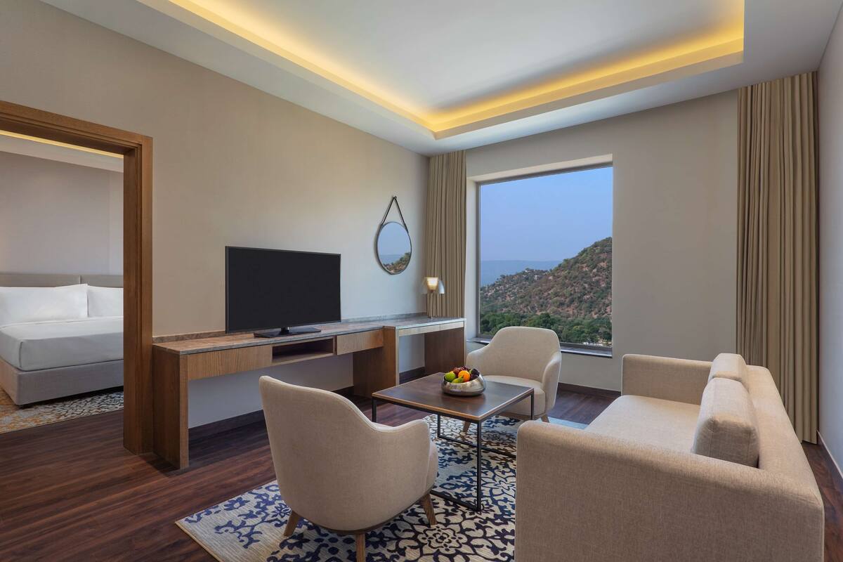 Una habitación de hotel moderna con un ventanal grande, un televisor de pantalla plana, un escritorio de madera, un sofá y una mesa de centro con fruta.