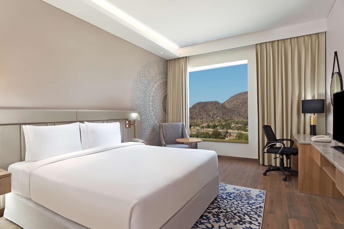 Habitación de hotel con una cama grande, un escritorio con silla, televisor y vista a un paisaje montañoso.