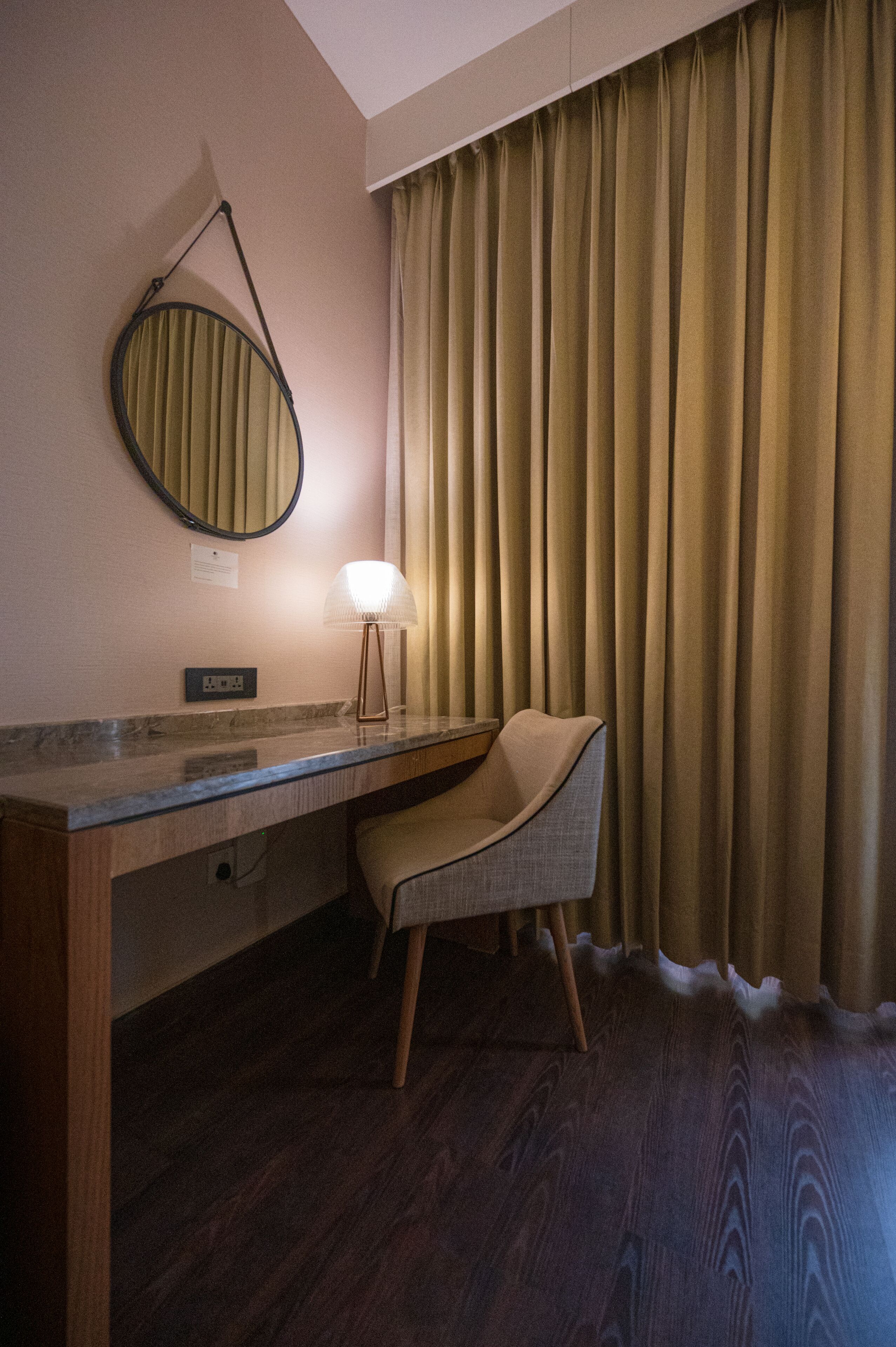 A hotel room with a wooden desk, a chair, a round mirror, a lamp, and a window with curtains.