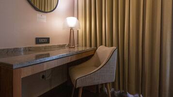 A hotel room with a wooden desk, a chair, a round mirror, a lamp, and a window with curtains.