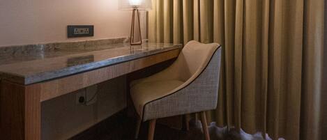 A hotel room with a wooden desk, a chair, a round mirror, a lamp, and a window with curtains.