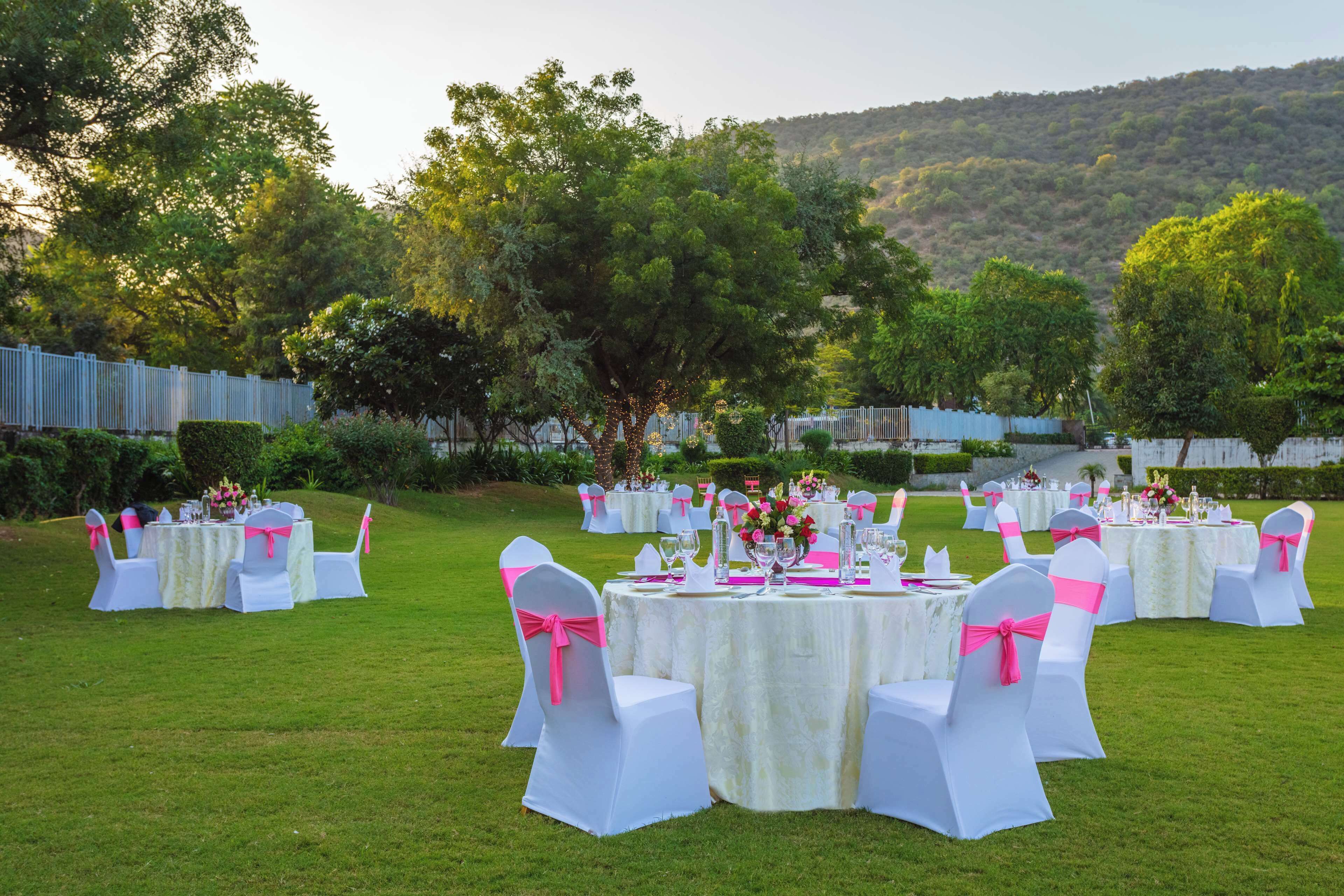 Un salón de bodas al aire libre con mesas redondas y sillas blancas, rodeado de follaje y colinas.
