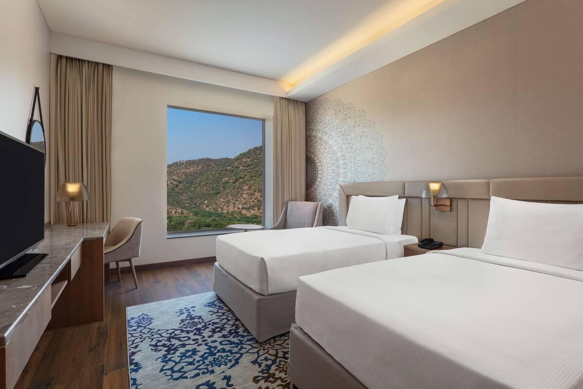 Habitación de hotel con dos camas, un escritorio, un televisor y un ventanal con vistas a un paisaje montañoso.