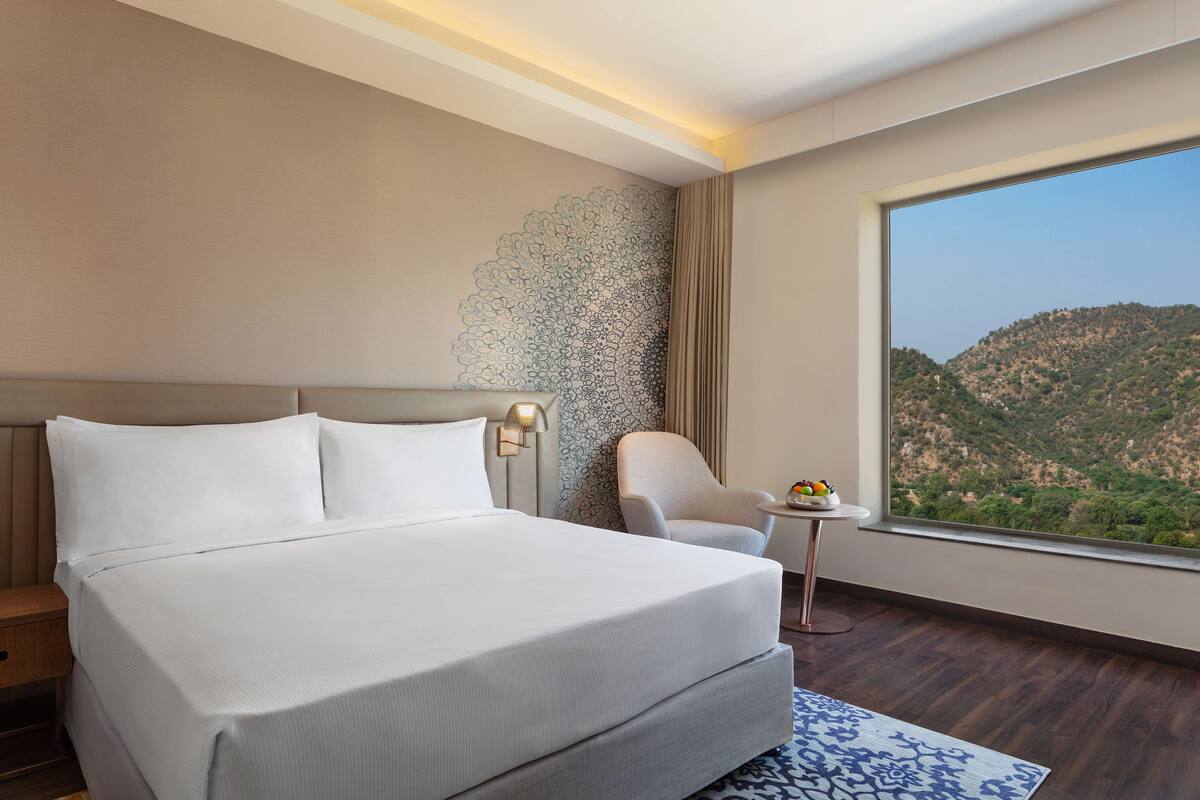 Habitación de hotel con una cama grande, una silla, una mesita y vistas a un paisaje montañoso.