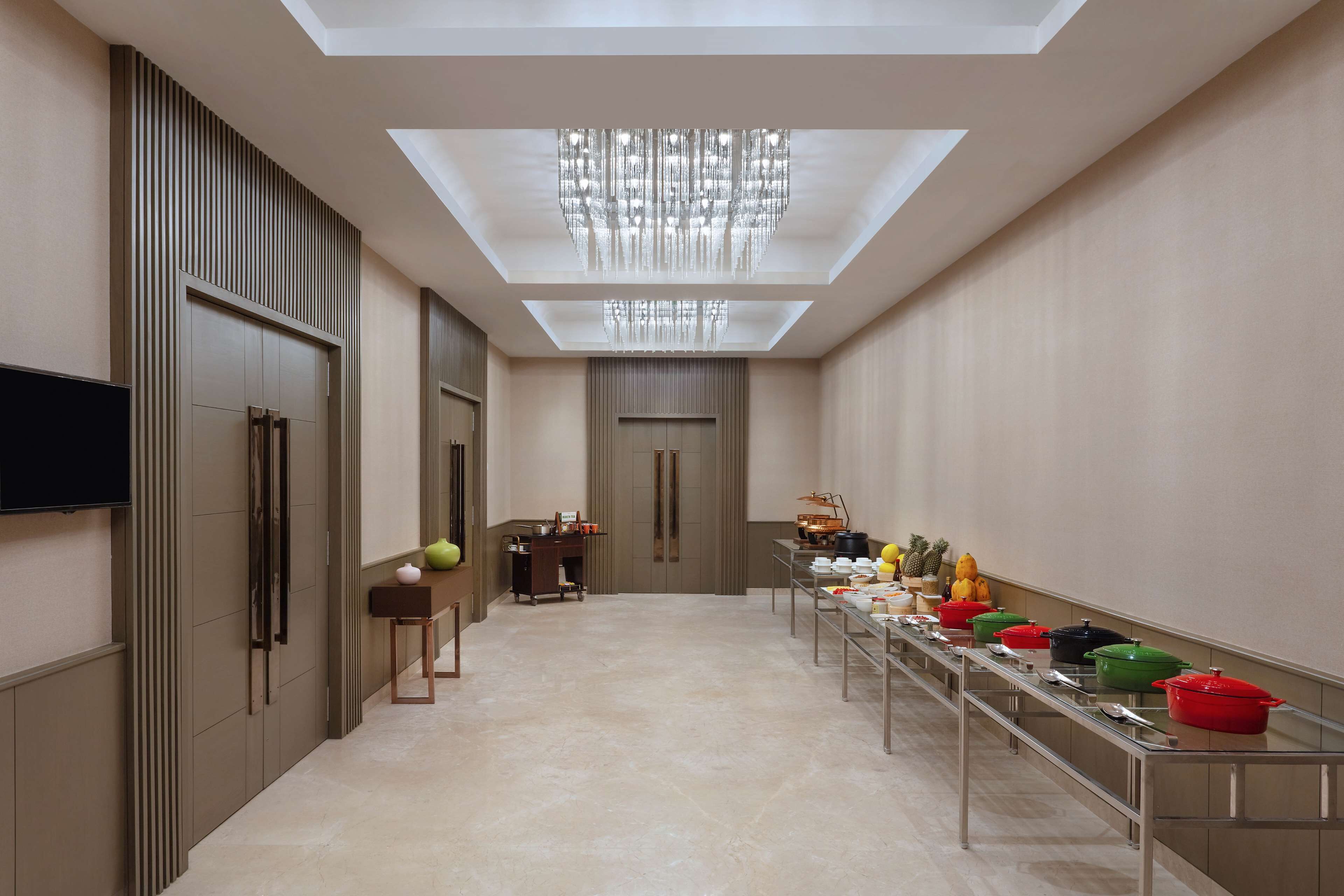 A modern hotel corridor with a long table displaying colorful pots, a flat-screen TV mounted on the wall, and a ceiling light fixture.