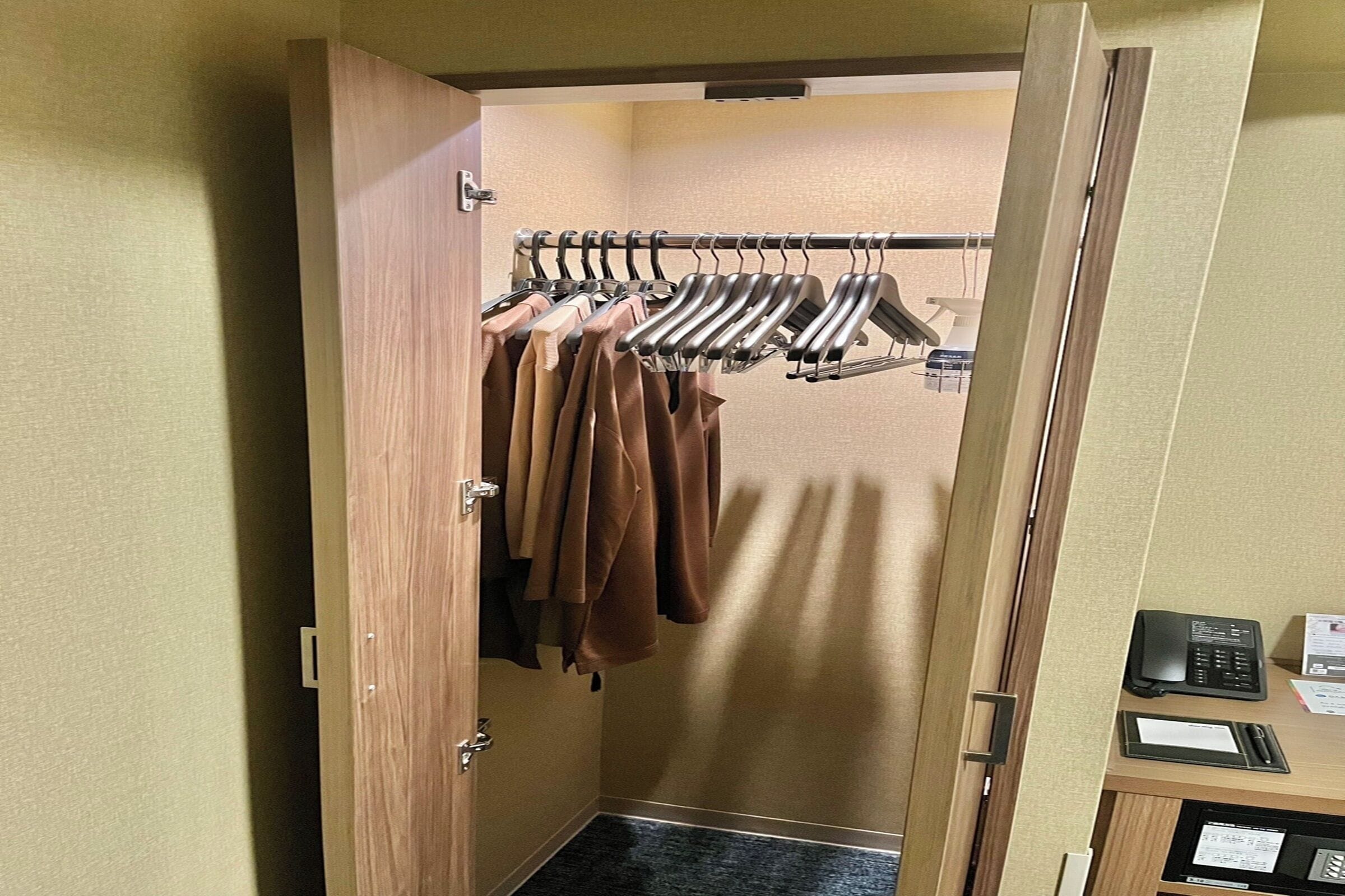 A hotel room with a wooden wardrobe, a coat rack with hanging clothes, a desk with a telephone and papers, and a small black object on the desk.