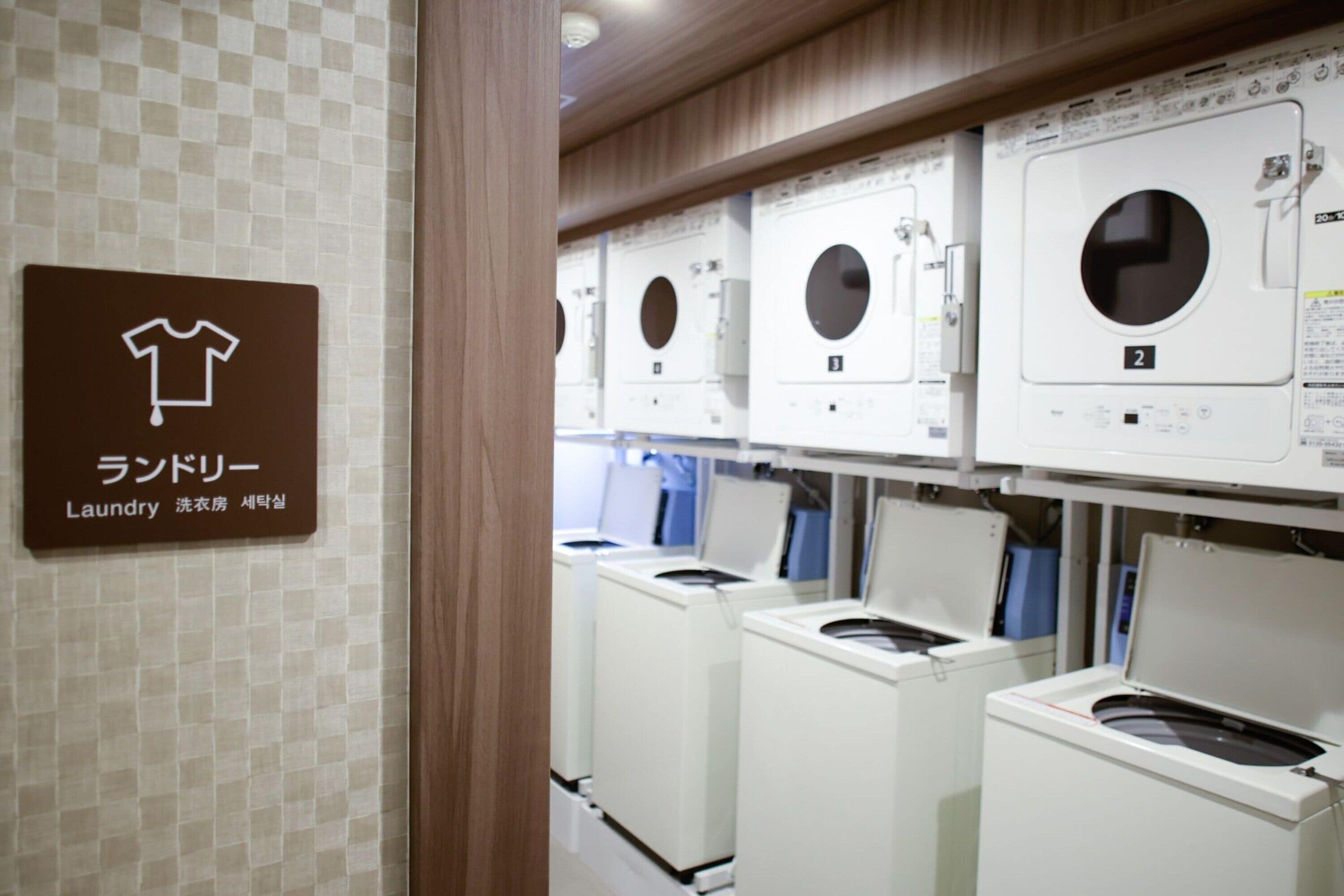 A laundry facility with multiple washing machines and dryers, a sign indicating the location, and a tiled wall.