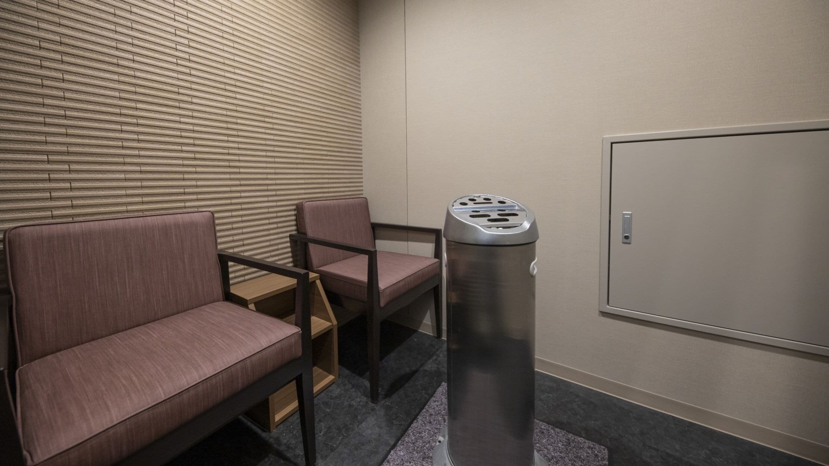 Habitación de hotel con zona de estar que incluye dos sillones acolchados y una mesita. Un cubo de basura está colocado cerca de la pared.