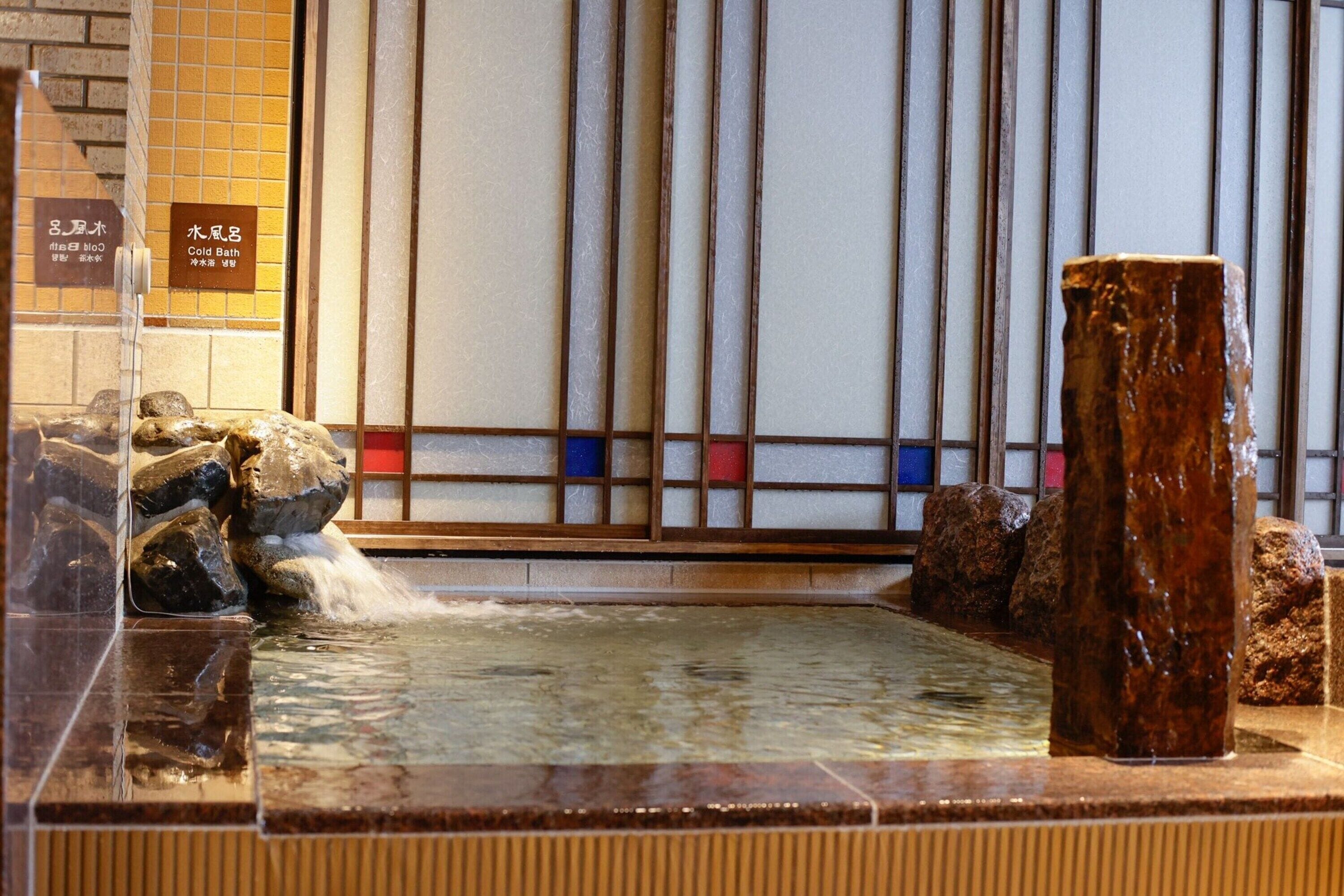 A traditional Japanese onsen with a stone bath, wooden pillars, and a tiled wall with a grid pattern.