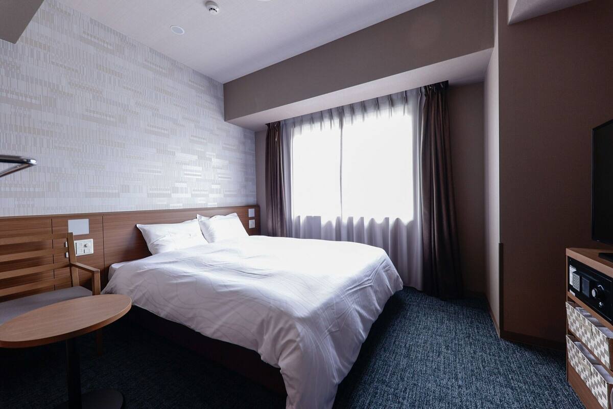 A hotel room with a large bed, a wooden headboard, a small round table, a television, and a window with curtains.