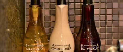 Trois bouteilles de produits Aroma Ess. Gold : shampoing, après-shampoing et savon pour le corps, posées sur une étagère carrelée.