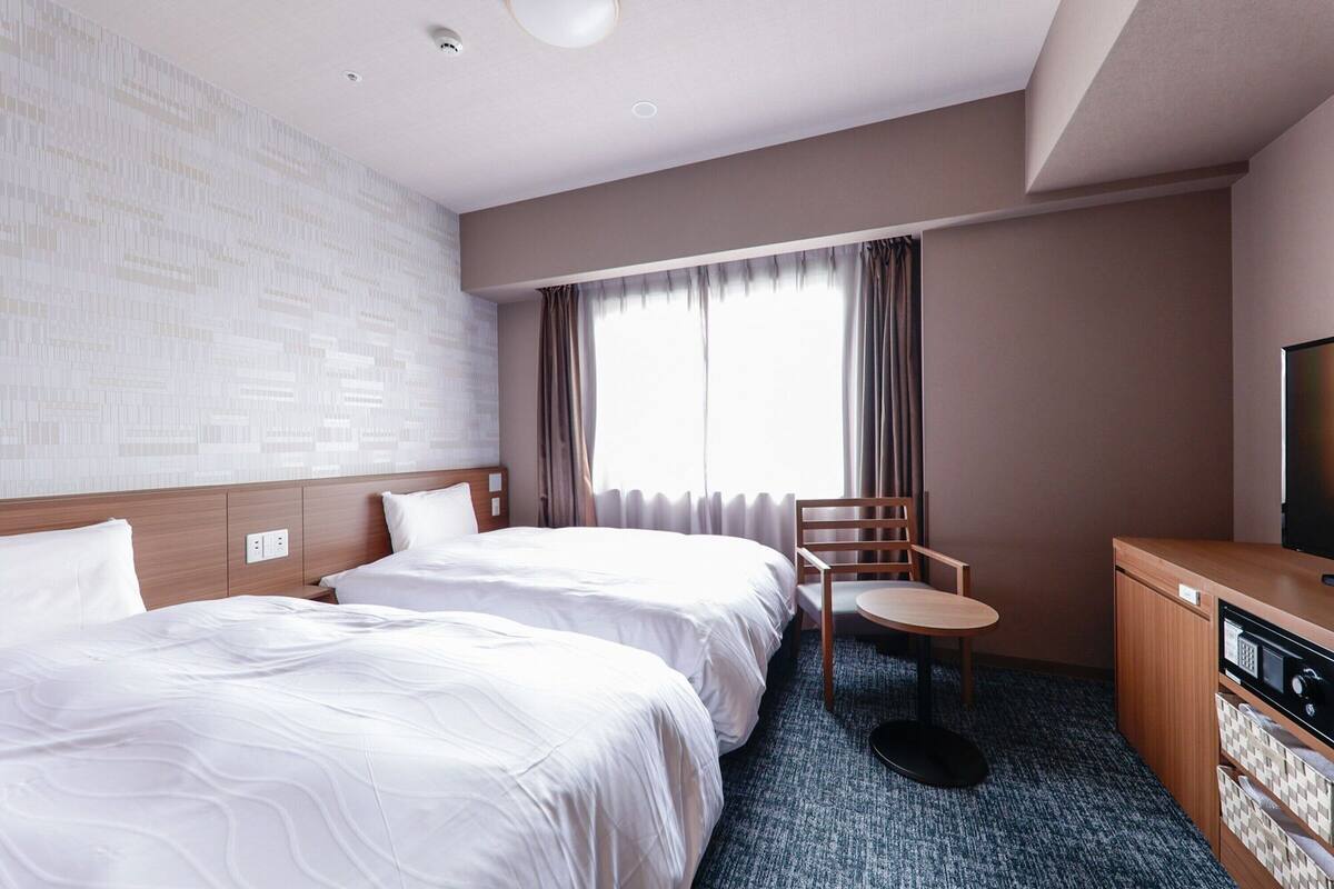 A hotel room with two beds, a small table, a chair, a TV, and a kitchenette.