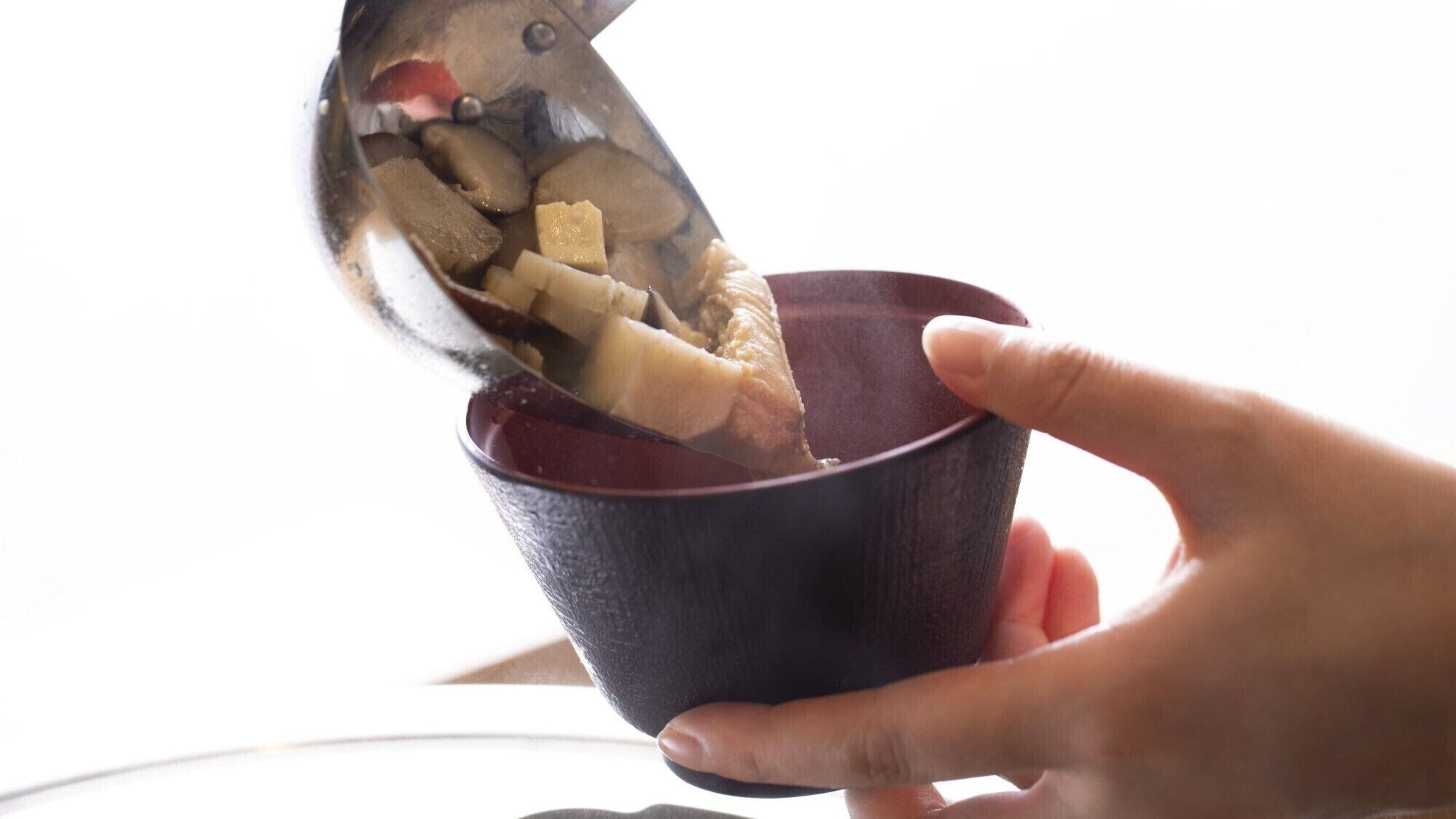 A hand holding a dark bowl with a spoon inside, containing a brownish liquid and some solid pieces.