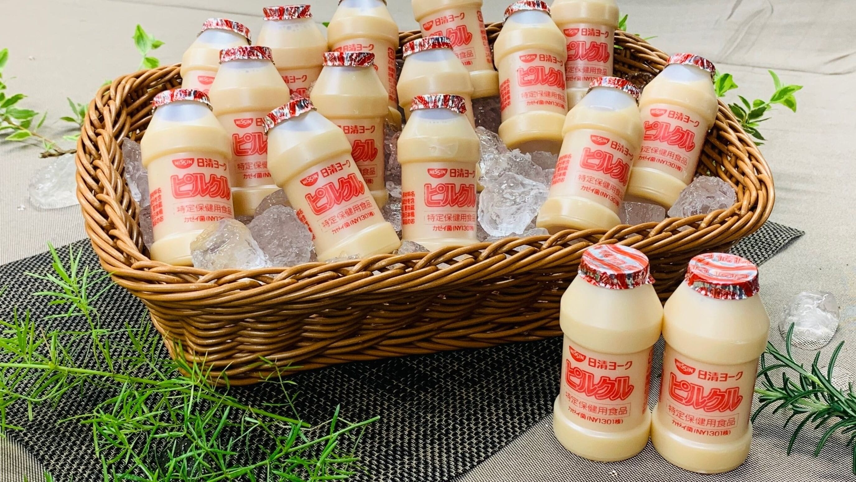 A wicker basket filled with bottles of a beverage, placed on a textured surface with greenery around.