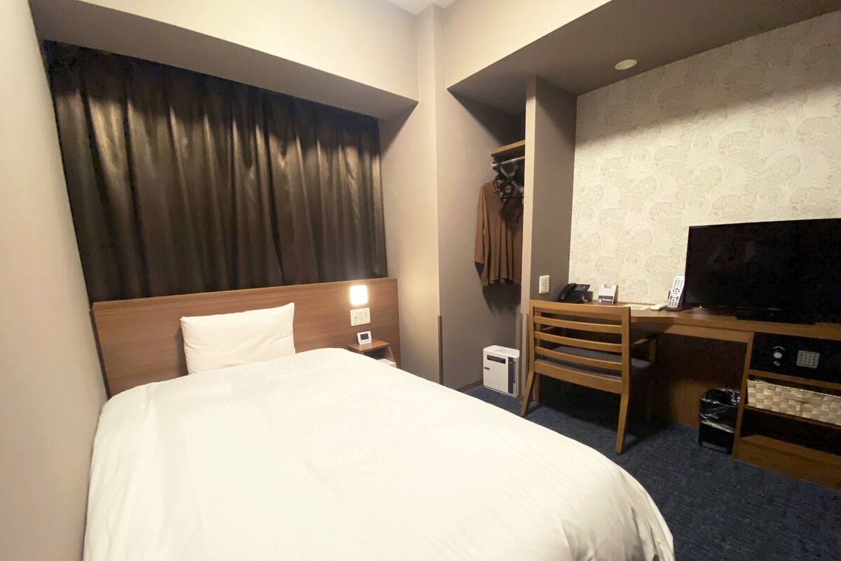 A hotel room with a bed, a desk with a television, a chair, and a coat rack.