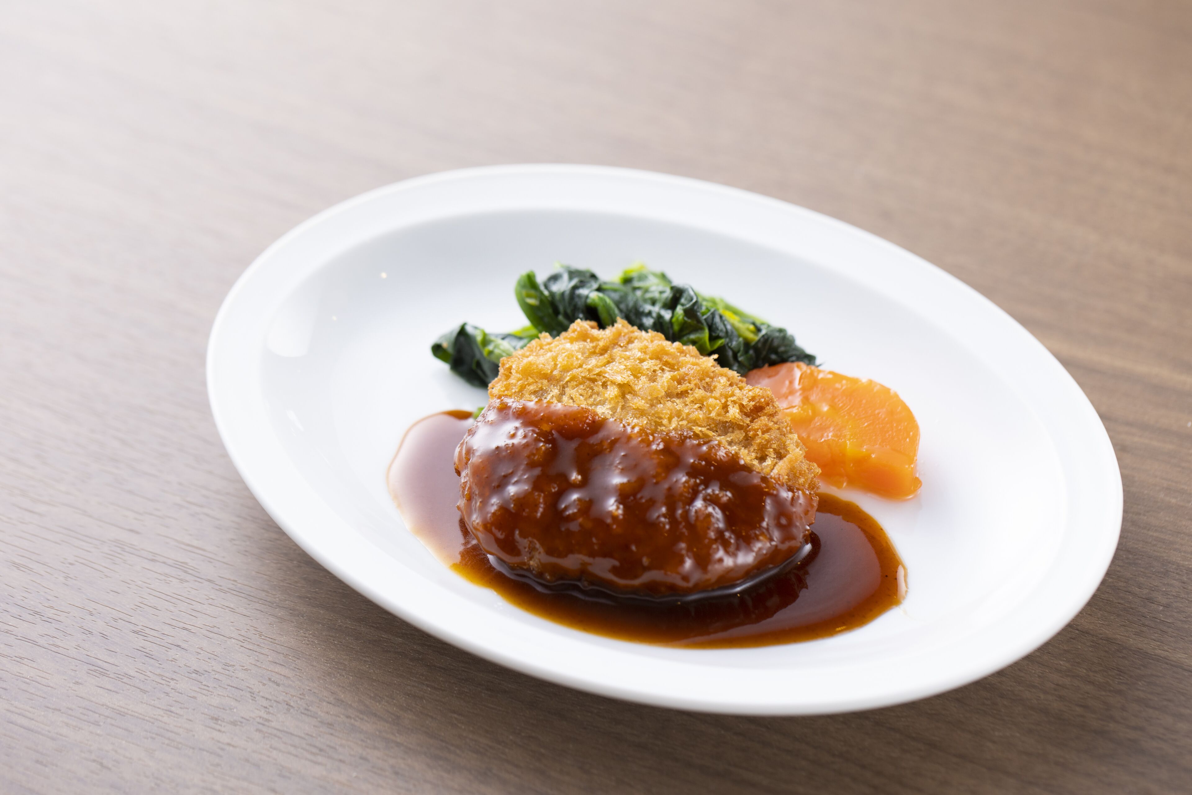 Un plato blanco con carne cubierta de pan rallado y salsa, acompañado de vegetales verdes y una rodaja de naranja.