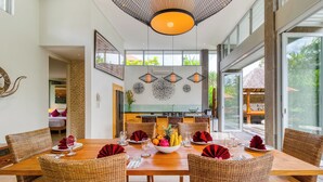 Een eethoek met een houten tafel, rieten stoelen en een fruitschaal. Er hangen hanglampen en in de achtergrond is een keuken te zien.