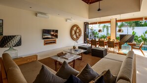 A spacious living area with a large sofa, coffee table, and a TV mounted on the wall. There is a dining area with chairs and a table, and a staircase leading to an outdoor area with plants. - Villa The Maya (Canggu)